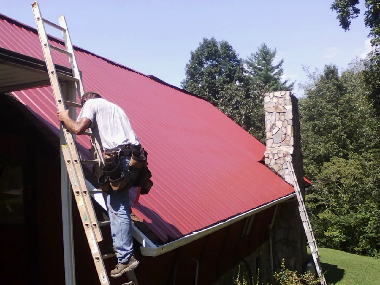Roofer on a ladder beside a red metal roof and stone chimney — Soffit & Fascia Repair by Roofing Brothers in Poquoson